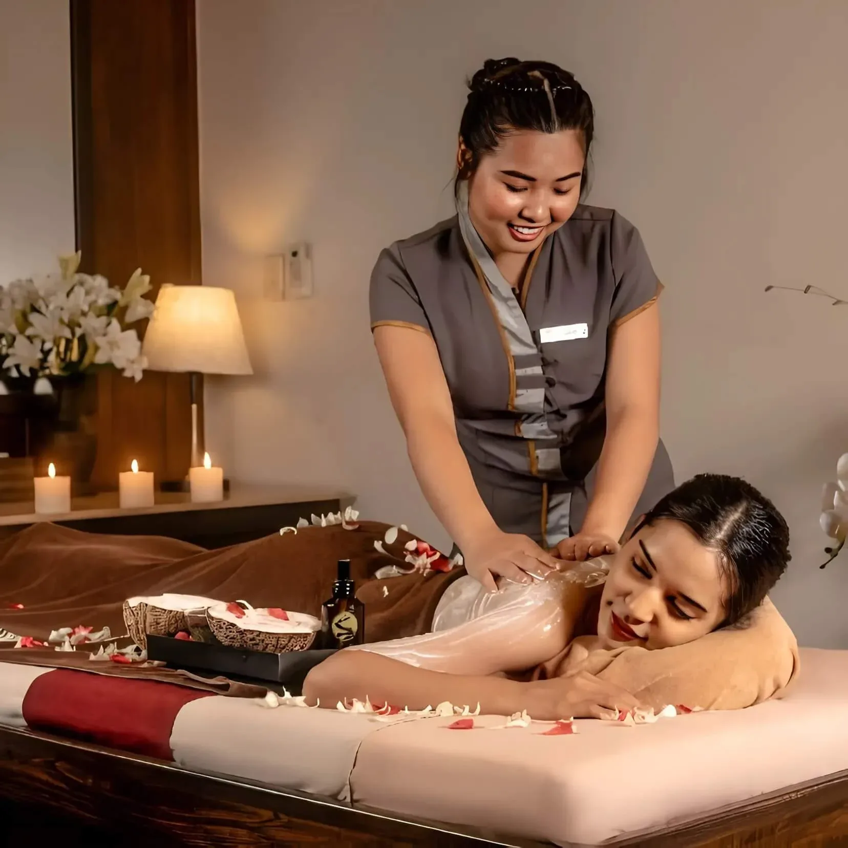 Smiling uniformed therapist applying a body treatment, lilies and candles on the table