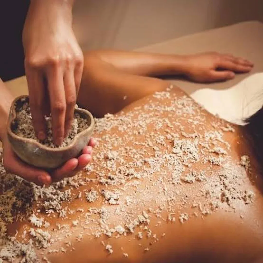 Hand scooping scrub from a stone bowl onto an oiled back under warm low light